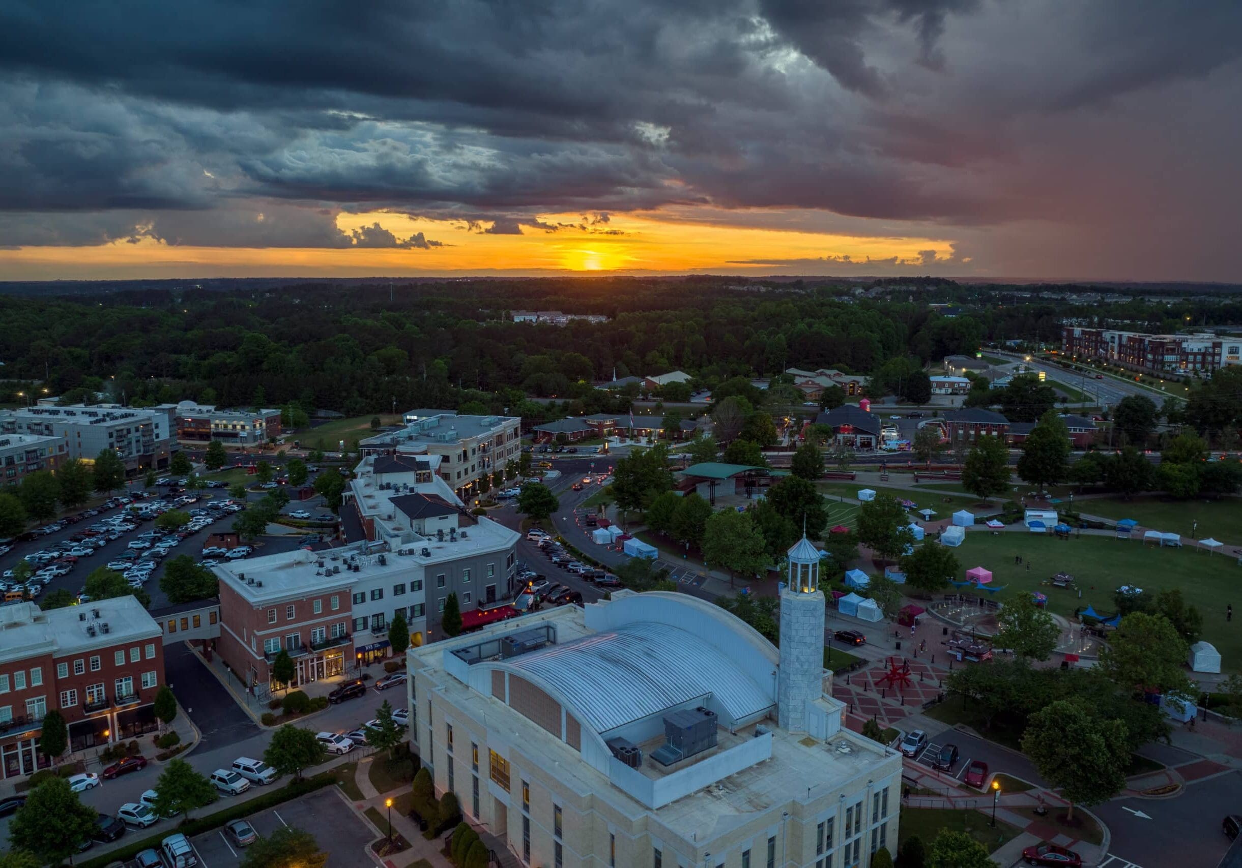 Aerial sunset view of Suwanee, Georgia, showing downtown buildings, green park, and dramatic clouds over the city landscape.